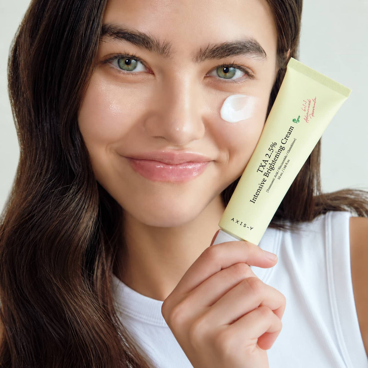 A woman holding a pale yellow tube of AXIS-Y TXA 2.5% Intensive Brightening Cream 50ml next to a dollop of white cream on her cheek, set against a clean, light background.