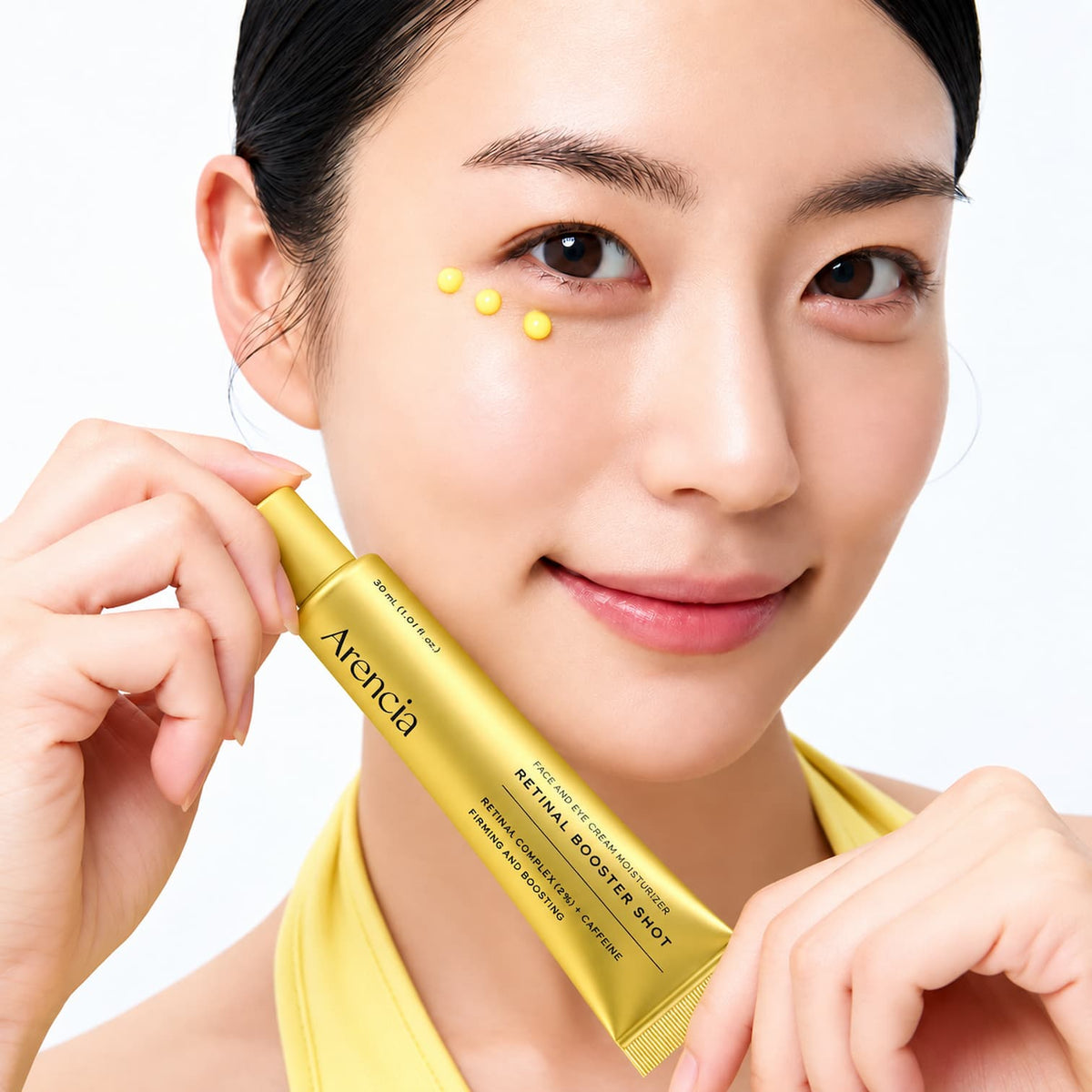 A woman holding an Arencia Retinal Booster Shot tube next to her cheek where three small yellow dots of the serum are applied, against a clean white background.