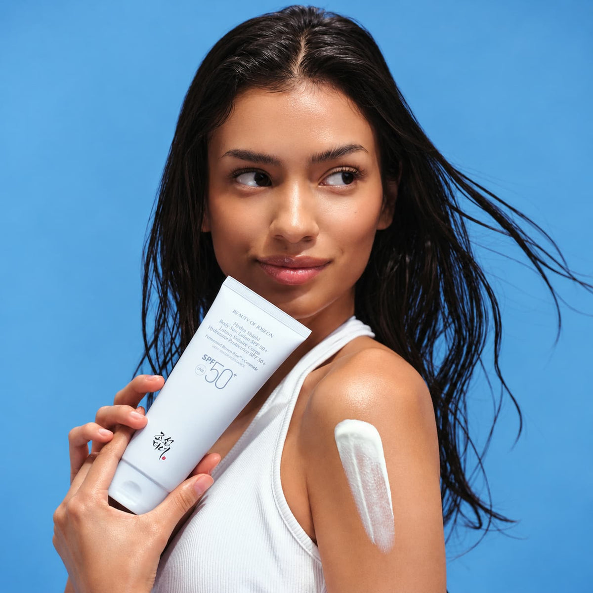 A woman with wet dark hair holds BEAUTY OF JOSEON Hydra Shield Body Sun Lotion against a bright blue background, with sunscreen visible on her shoulder.