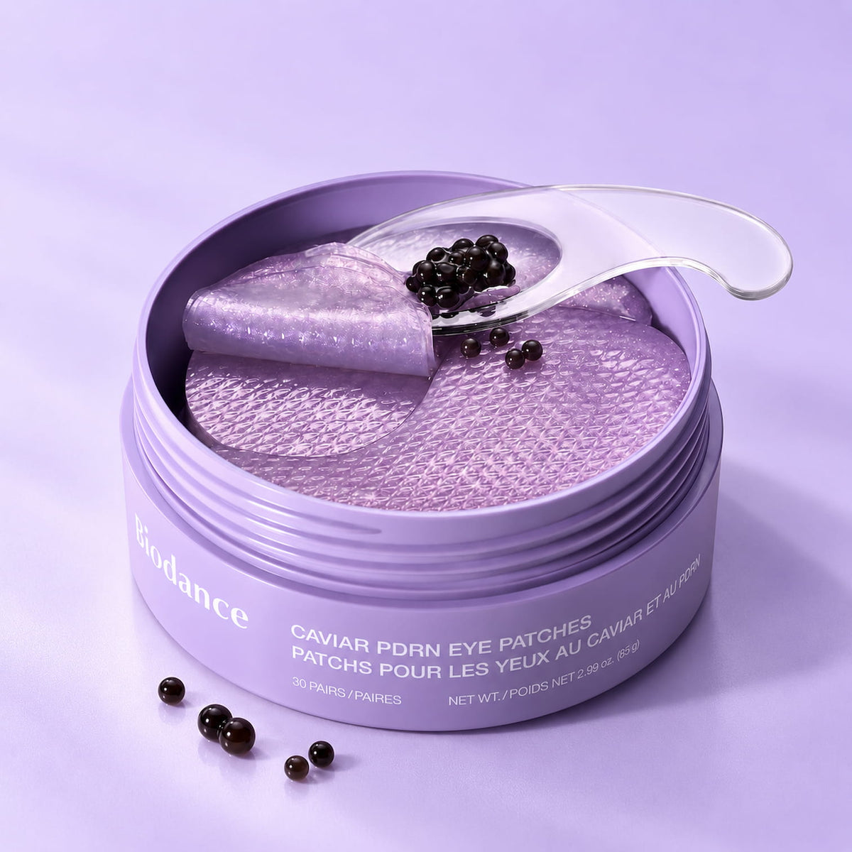 A round lavender cosmetic jar BIODANCE Caviar PDRN Eye Patches 30pcs containing shimmering lavender eye patches, with a clear spatula resting across the open lid and a few black caviar-like beads scattered on the product and surface.