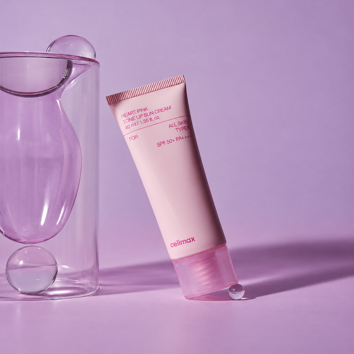 A tube of CELIMAX Heart Pink Tone Up Sun Cream 40ml in a pale pink color, standing upright on a lavender surface with a matching glass vessel on the left.