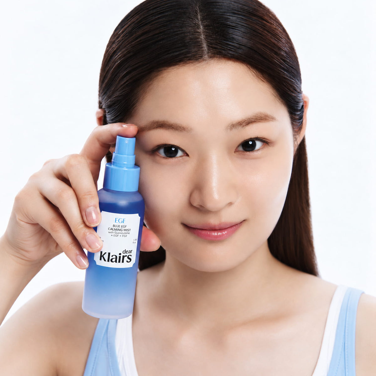 A woman holding a bottle of DEAR KLAIRS Blue EGF Calming Mist 100ml next to her face against a clean white background.