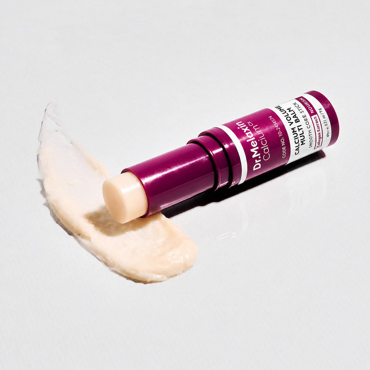 Dr. Melaxin Calcium volume multi balm stick with a deep magenta-purple cap, shown with a smear of the beige cream product on a light grey background.