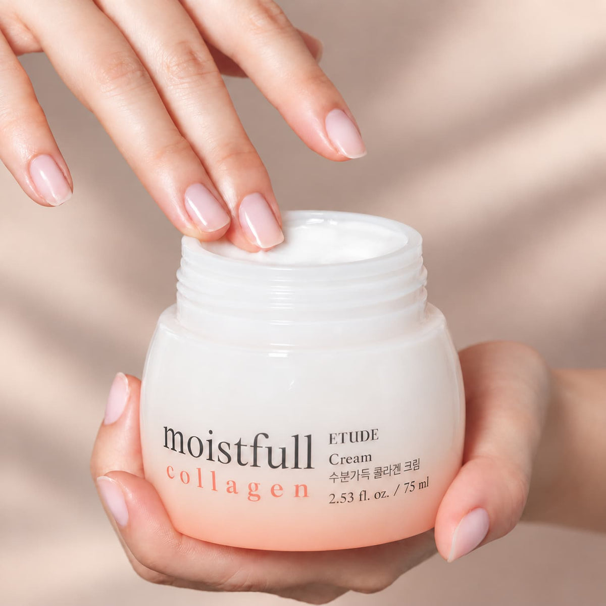 A close-up of a person holding an open jar of ETUDE Moistfull Collagen Cream, with their fingers resting on the white cream inside. The background is a soft, warm beige with elegant shadows.