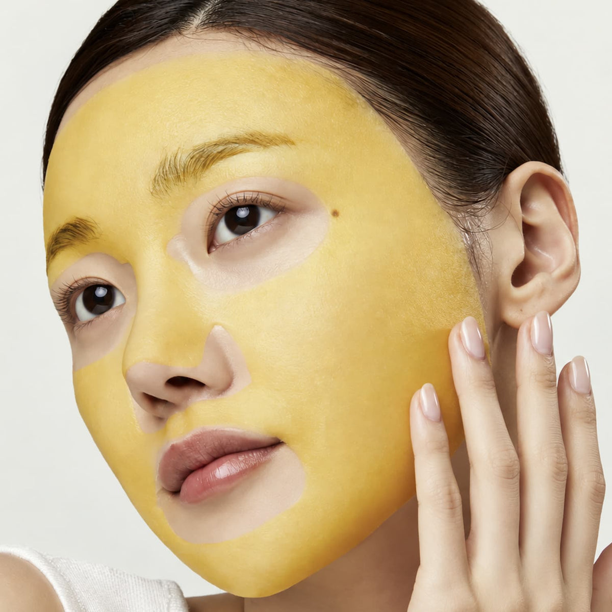 Close-up of a woman with a bright yellow skincare mask applied to her face, touching her cheek, against a light neutral background.