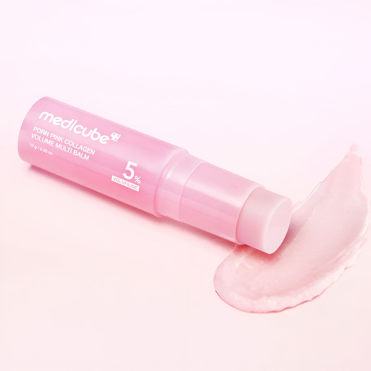 A pink tube of MEDICUBE PDRN Pink Collagen Volume Multi Balm 10g lies diagonally on a light pink background with a smear of the balm in front of it.