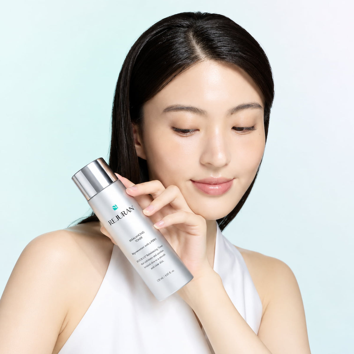 A woman with dark hair holds a bottle of REJURAN Rebalancing Toner 120ml on a light grey background.
