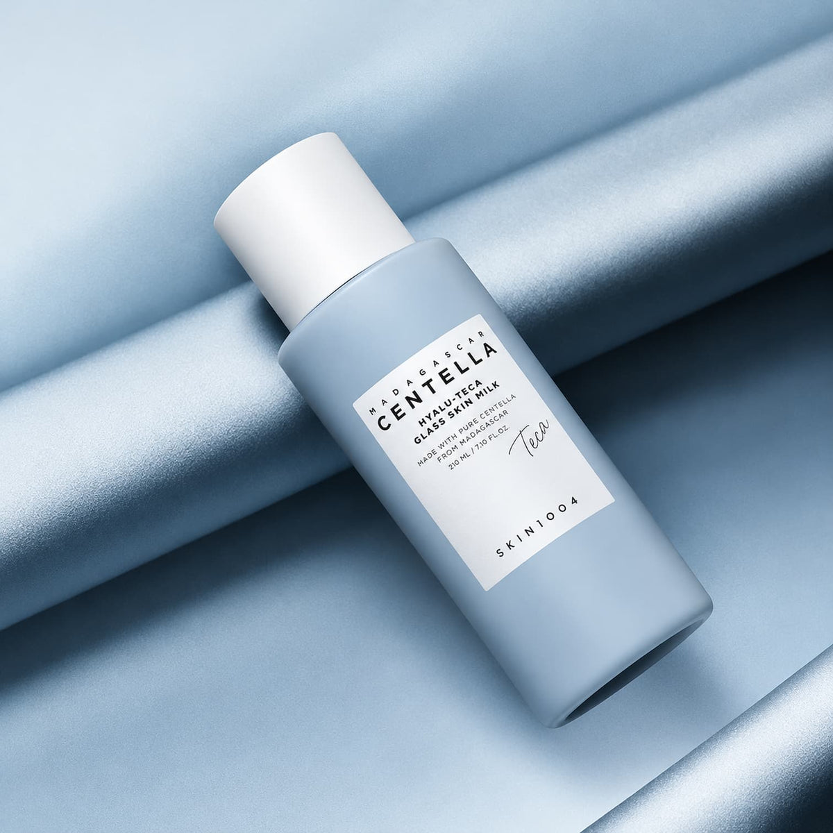 A bottle of SKIN1004 Madagascar Centella Hyalu-Teca Glass Skin Milk featuring a light blue-gray bottle and white cap, resting on a draped light blue satin fabric.
