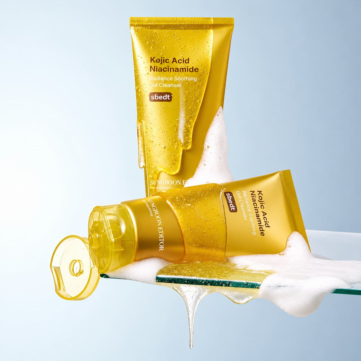 Two gold tubes of SUNGBOON EDITOR Kojic Acid Niacinamide Radiance Soothing Gel Cleanser, one standing upright and one lying on its side, both covered in water droplets and dripping white foam, set against a light blue gradient background.