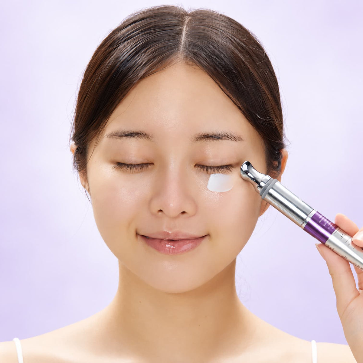A close-up of a woman with her eyes closed as she uses a silver and purple eye cream roller tube on her under-eye area, with white cream visible on her cheek against a soft lavender background.