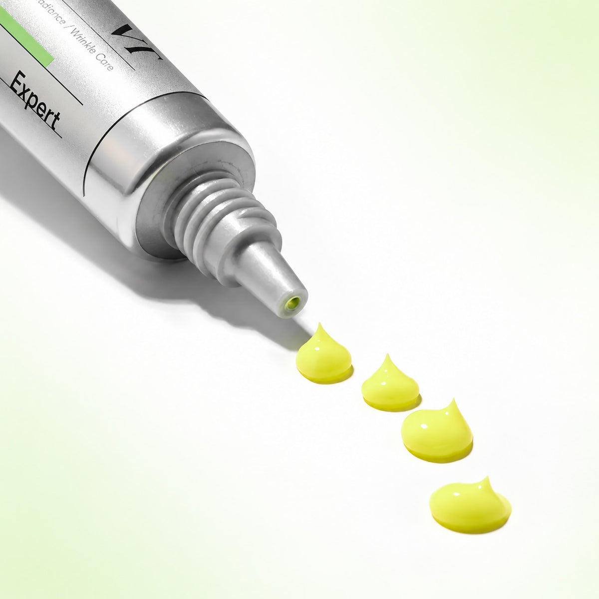 A silver cosmetic tube labelled 'Expert' lies diagonally on a white, green background, with four small dollops of bright yellow cream or gel dripping from the tip in a row.