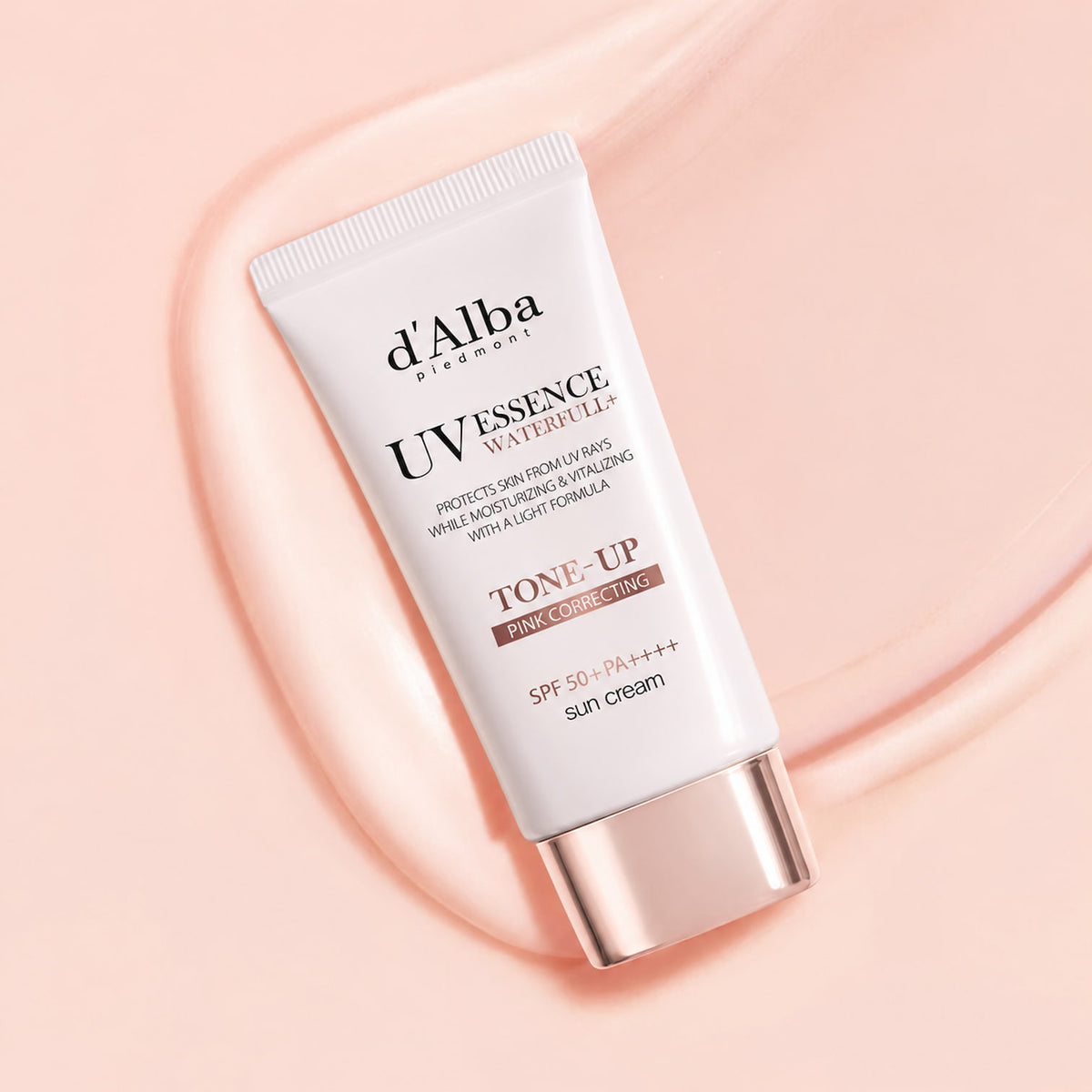 A white squeeze tube of d'Alba UV Essence pink correcting Tone-Up Sun Cream with a metallic rose gold cap, set against a matching peach-pink background with a wavy texture.