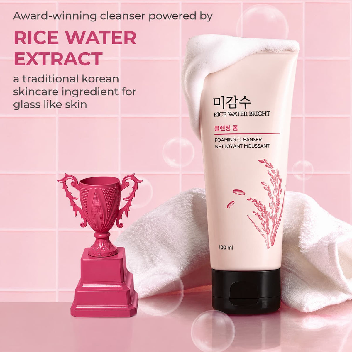 A 100 ml tube of Rice Water Bright Foaming Cleanser standing upright on a pink reflective surface next to a pink trophy and a white towel. The background is a pink tiled wall with floating bubbles, and promotional text is visible on the left.