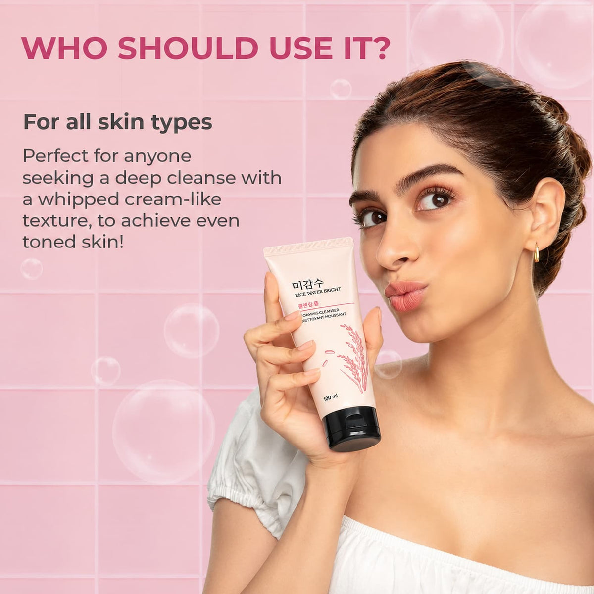 A woman holding a tube of facial cleanser against a pink tiled background with promotional text reading 'WHO SHOULD USE IT?'. The product tube is light pink with a black cap and Korean text on the front.